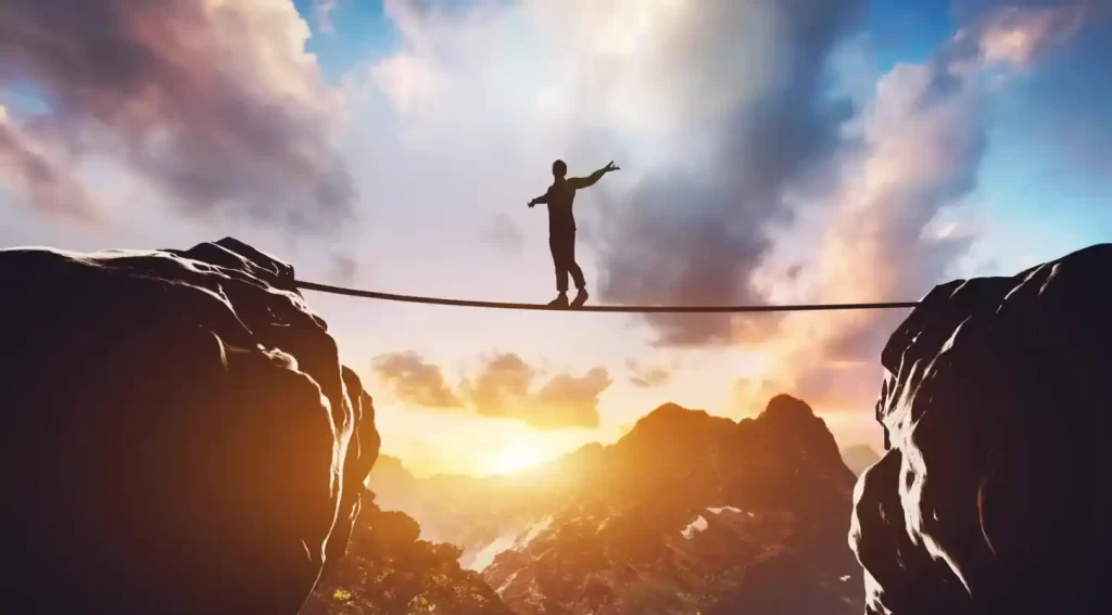 silhouette of a man with outstretched arms walking a tightrope between two cliffs with the sunset in the background