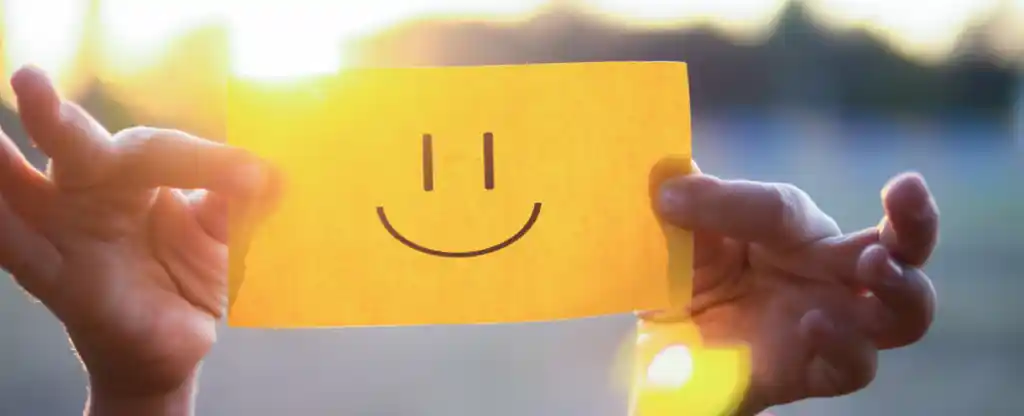 two hands holding a post it note with a smiley face drawn on and sunlight in the background