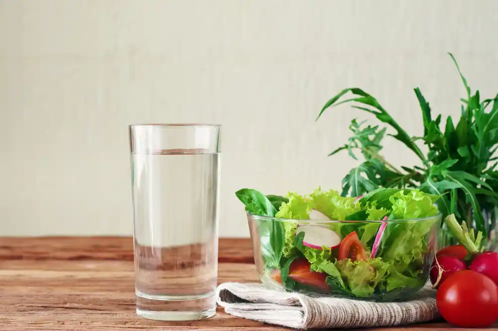glass of water on a table next to a bowl of salad and tomatoes