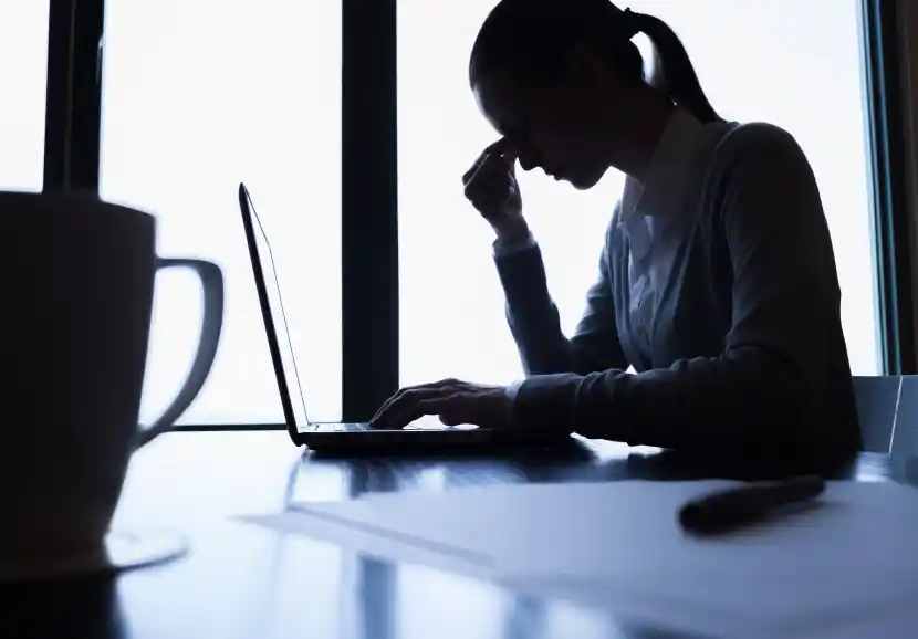 silhouette of woman sitting behind her laptop pinching the top of her nose between the eyes