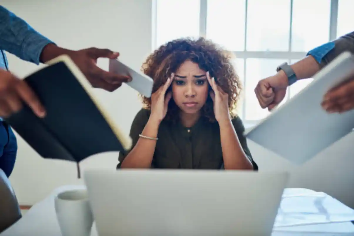 woman sitting behind a laptop looking overwhelmed and hands around her trying to add more tasks